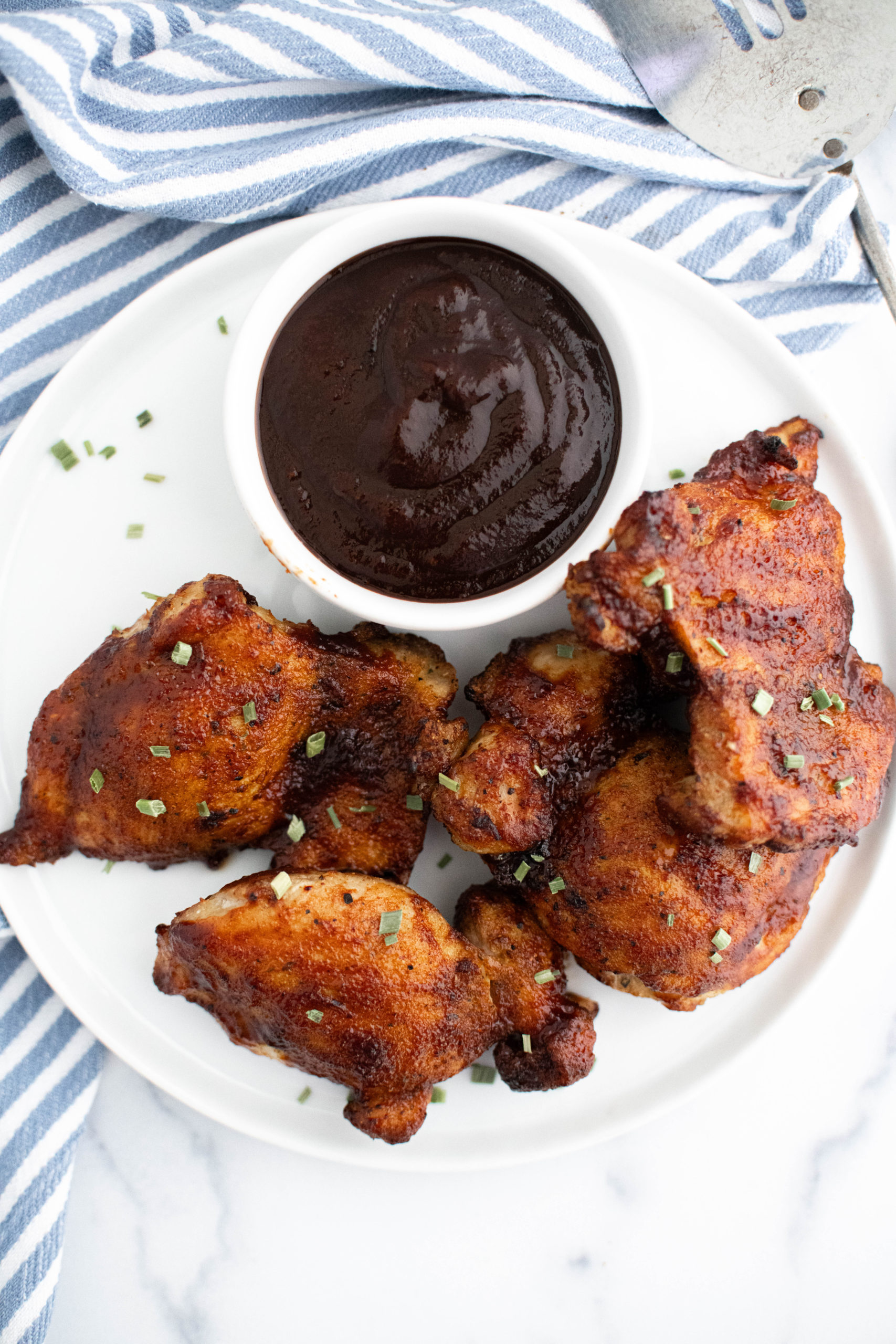 Straight down shot of a white plate with barbecue chicken thighs and a small bowl of barbecue sauce.