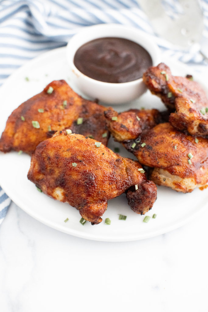 Barbecue chicken thighs on a round white plate with a small bowl of barbecue sauce on the plate.