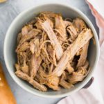 Shredded slow cooker Italian pork in a gray bowl. Bottle of dressing in upper left corner and baguette in lower left corner. Pale pink cloth napkin draped on right side of photo.