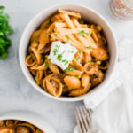 One bowl of taco pasta in center of photo. Another bowl in lower left corner. Cilantro and taco seasoning in background.