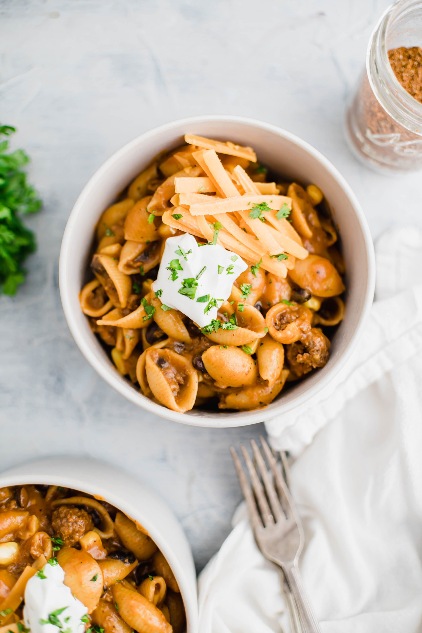 One bowl of taco pasta in center of photo. Another bowl in lower left corner. Cilantro and taco seasoning in background.