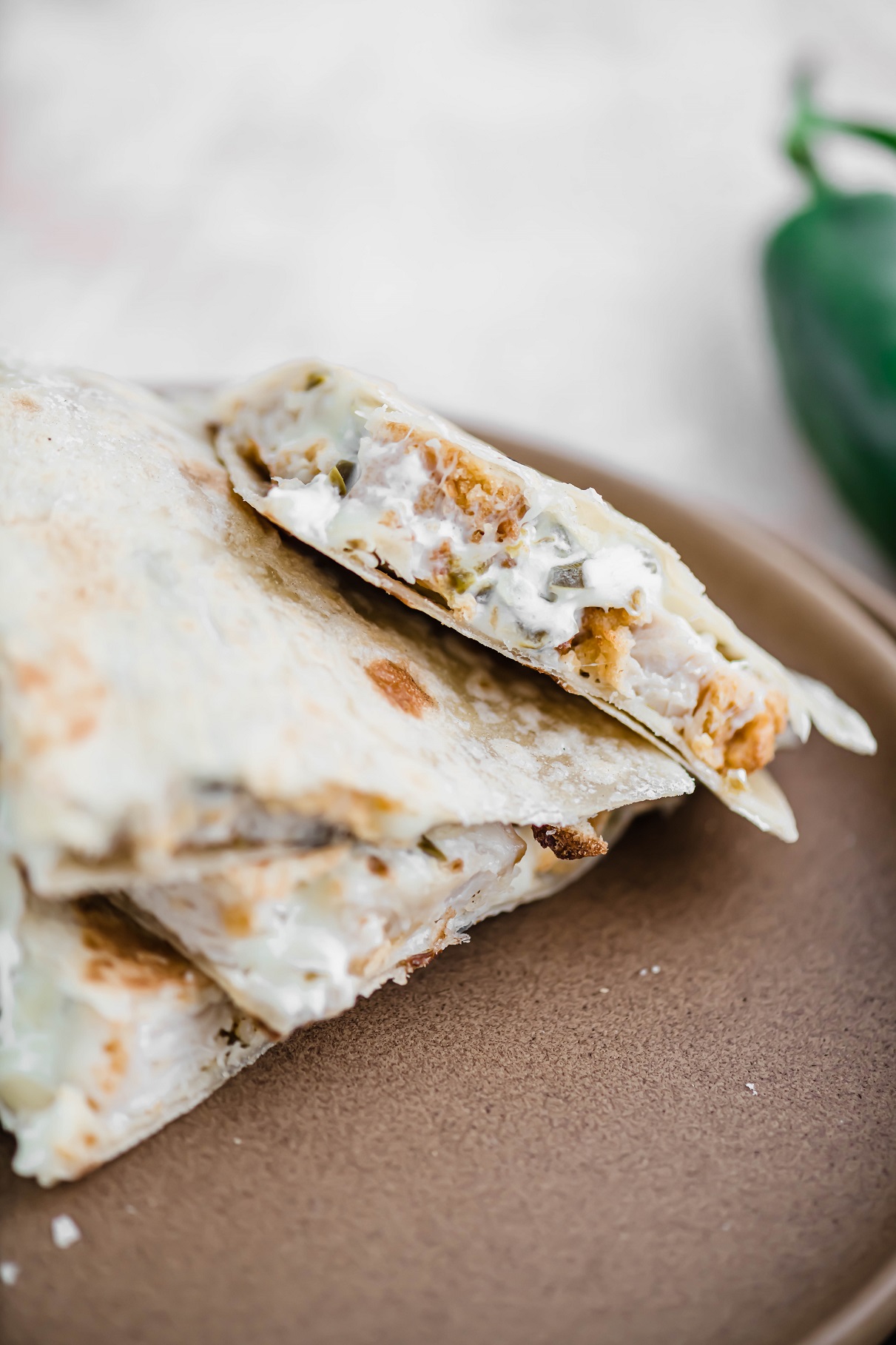 Sliced jalapeno popper quesadilla on a clay colored plate, exposing the inside of the quesadilla. Jalapeno in the background.