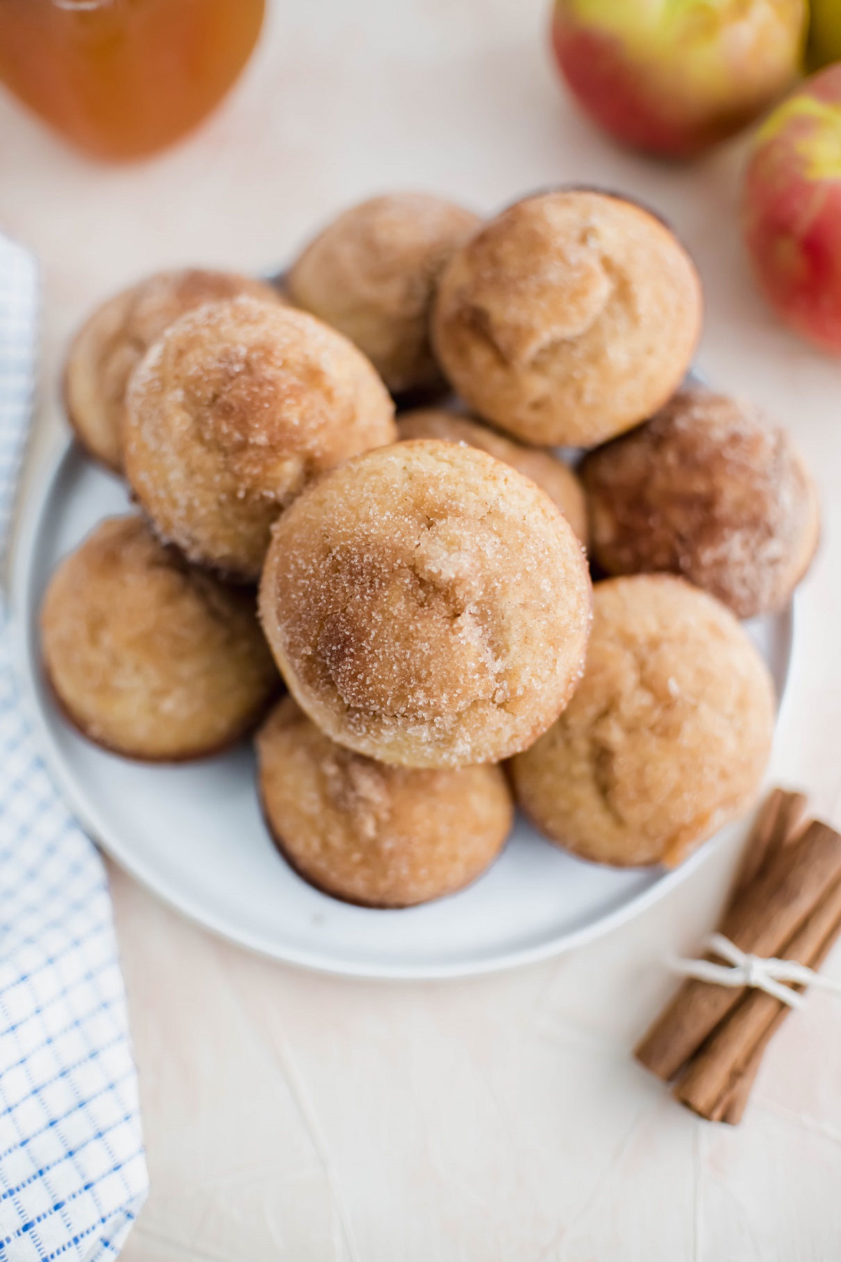 Dozen apple cider muffins piled on a round white plate with a tied bundle of cinnamon sticks in lover right corner and apples and apple cider in background.