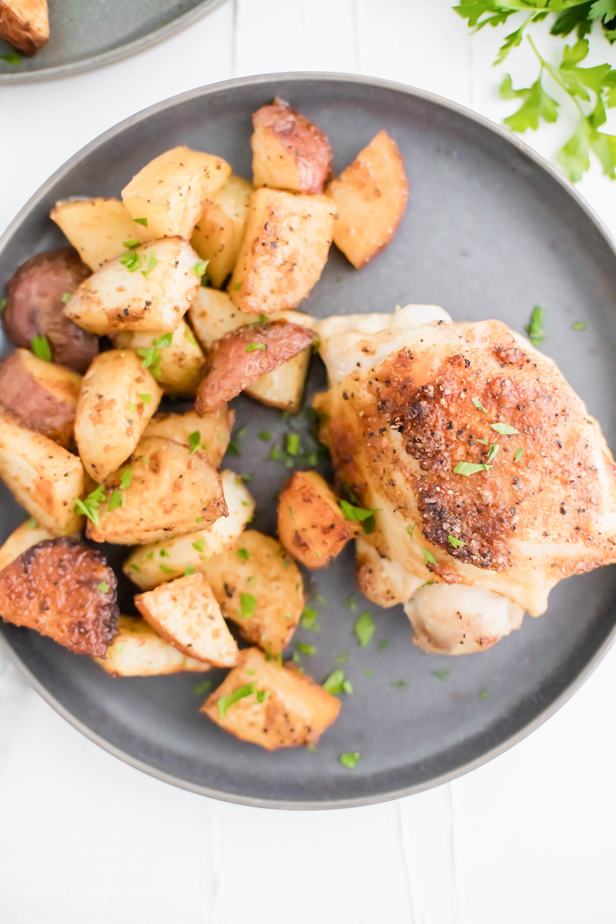 Baked chicken and potatoes on a round gray plate with sprigs of parsley in the upper right corner.