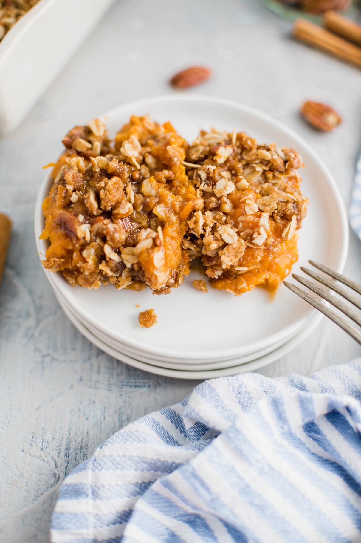 Two scoops of sweet potato crunch on a stack of small round white plates. Fork leaning on the edg of the plate with a blue and white striped cloth napkin in lower right corner.