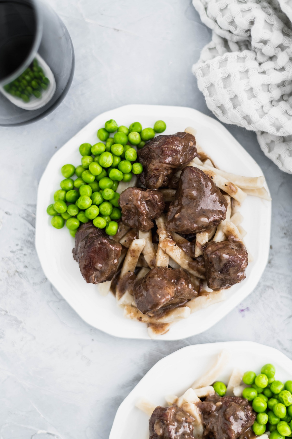 Tender instant pot beef tips over egg noodles with a side of peas on a small white round plate. Another prepared plate peeking in lower right corner.