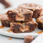 Individual rolo brownie sliced in half to show melted rolo inside. Set on a white round plate with more brownie bites in the background.