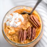 Straight down photo of a jar of pumpkin overnight oats garnished with three pecan halves, a dollop of whipped cream and a sprinkle of cinnamon. Jar has a spoon in it with a cloth napkin draped on the right side of the jar.