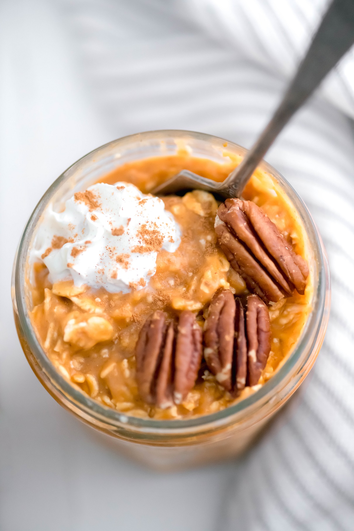Straight down photo of a jar of pumpkin overnight oats garnished with three pecan halves, a dollop of whipped cream and a sprinkle of cinnamon. Jar has a spoon in it with a cloth napkin draped on the right side of the jar.