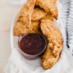 White plastic food basket with parchment paper, filled with air fryer chicken tenders and a small glass bowl of barbecue sauce.