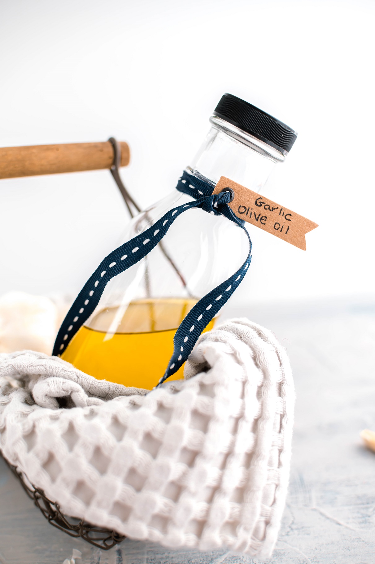 Glass bottle of garlic infused olive oil in a basket lined with a kitchen towel. Bottle tied with ribbon and a tag that states "garlic olive oil."