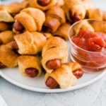 White round plate filled with air fryer pigs in a blanket with a glass bowl of ketchup on the plate.