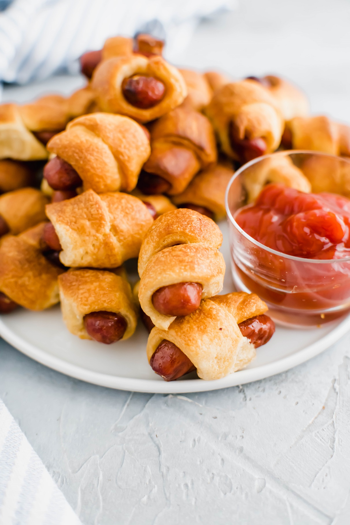 White round plate filled with air fryer pigs in a blanket with a glass bowl of ketchup on the plate.