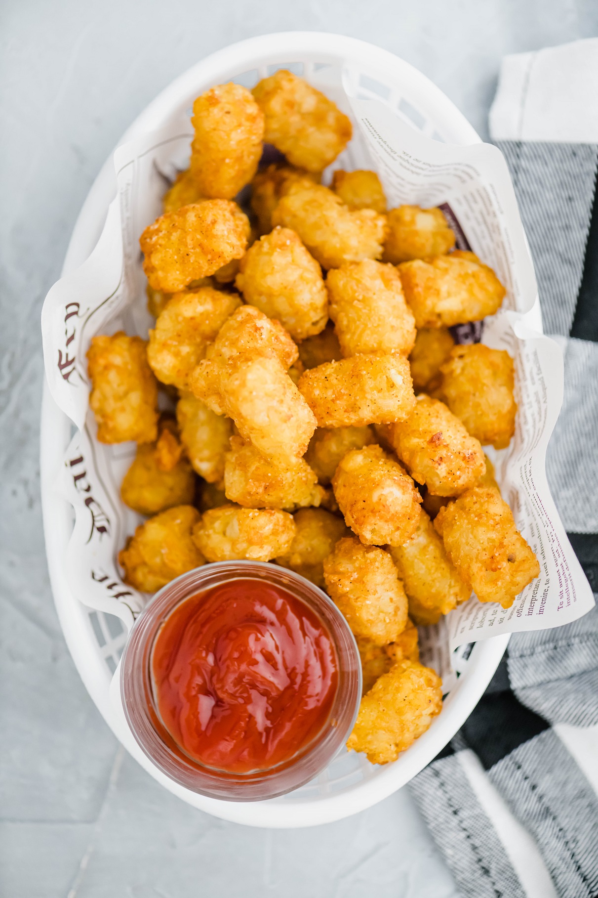 White plastic basket lined with newspaper parchment and filled with crispy tater tots and a small glass bowl of ketchup