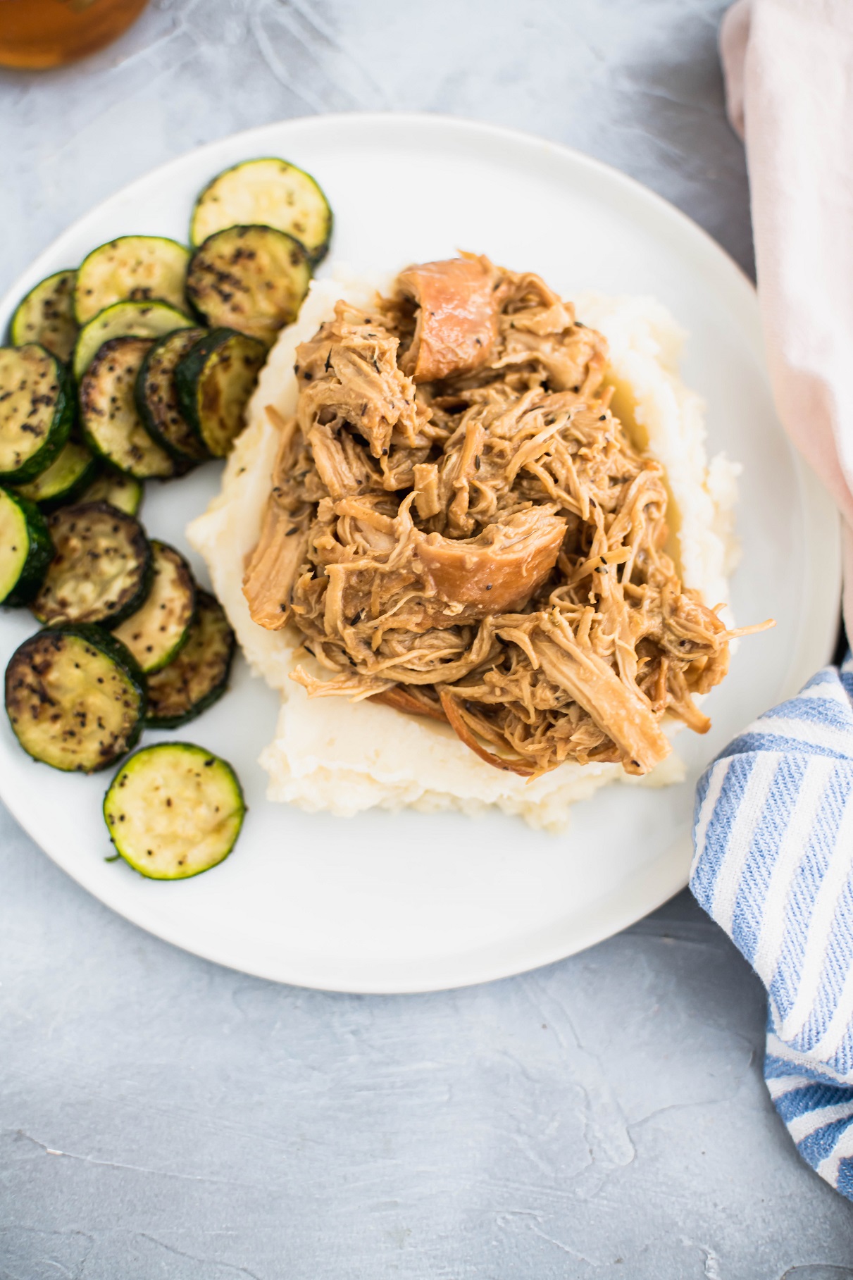 Round white plate with shredded honey mustard chicken on top of mashed potatoes with a side of sauteed zucchini.