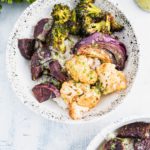 White bowl with black speckles filled with white rice that is topped with roasted broccoli, cauliflower, purple sweet potato chunks and a quarter of a red onion. Drizzled with sauce.