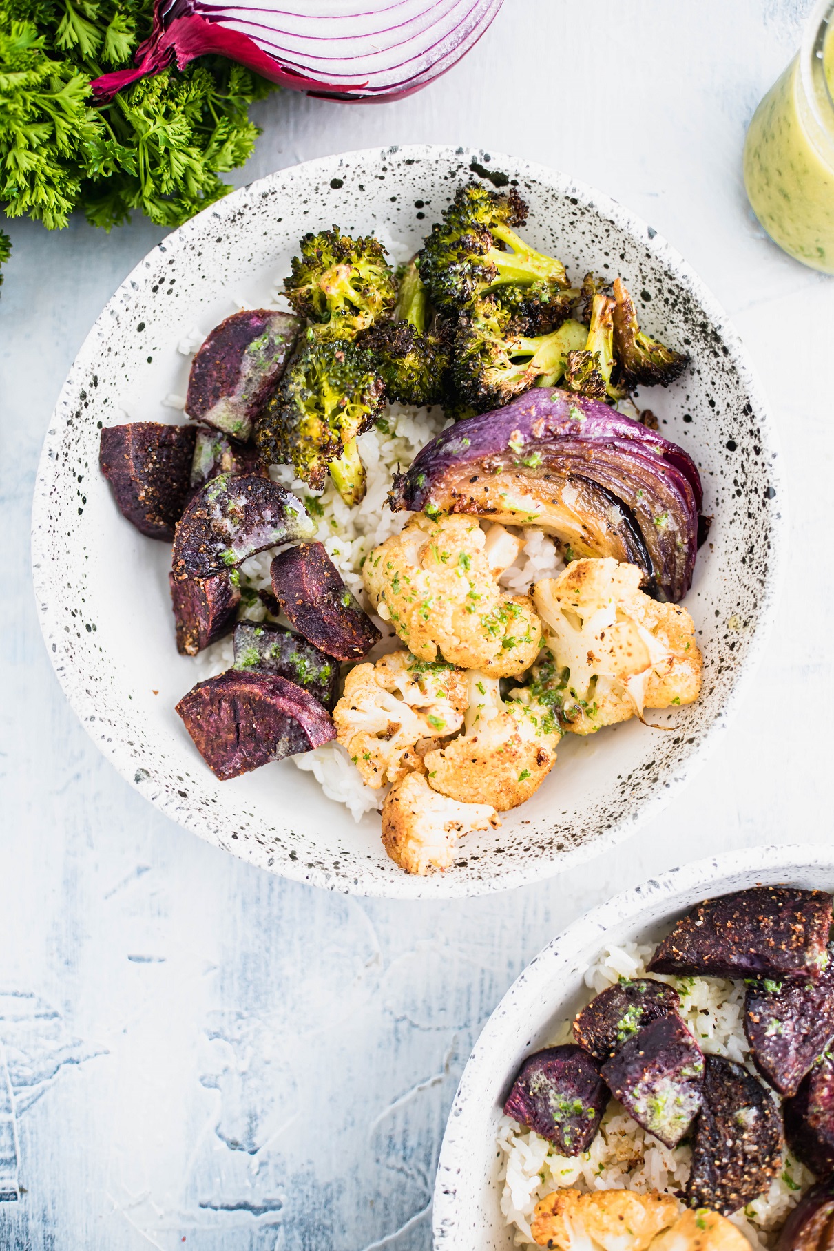 White bowl with black speckles filled with white rice that is topped with roasted broccoli, cauliflower, purple sweet potato chunks and a quarter of a red onion. Drizzled with sauce.
