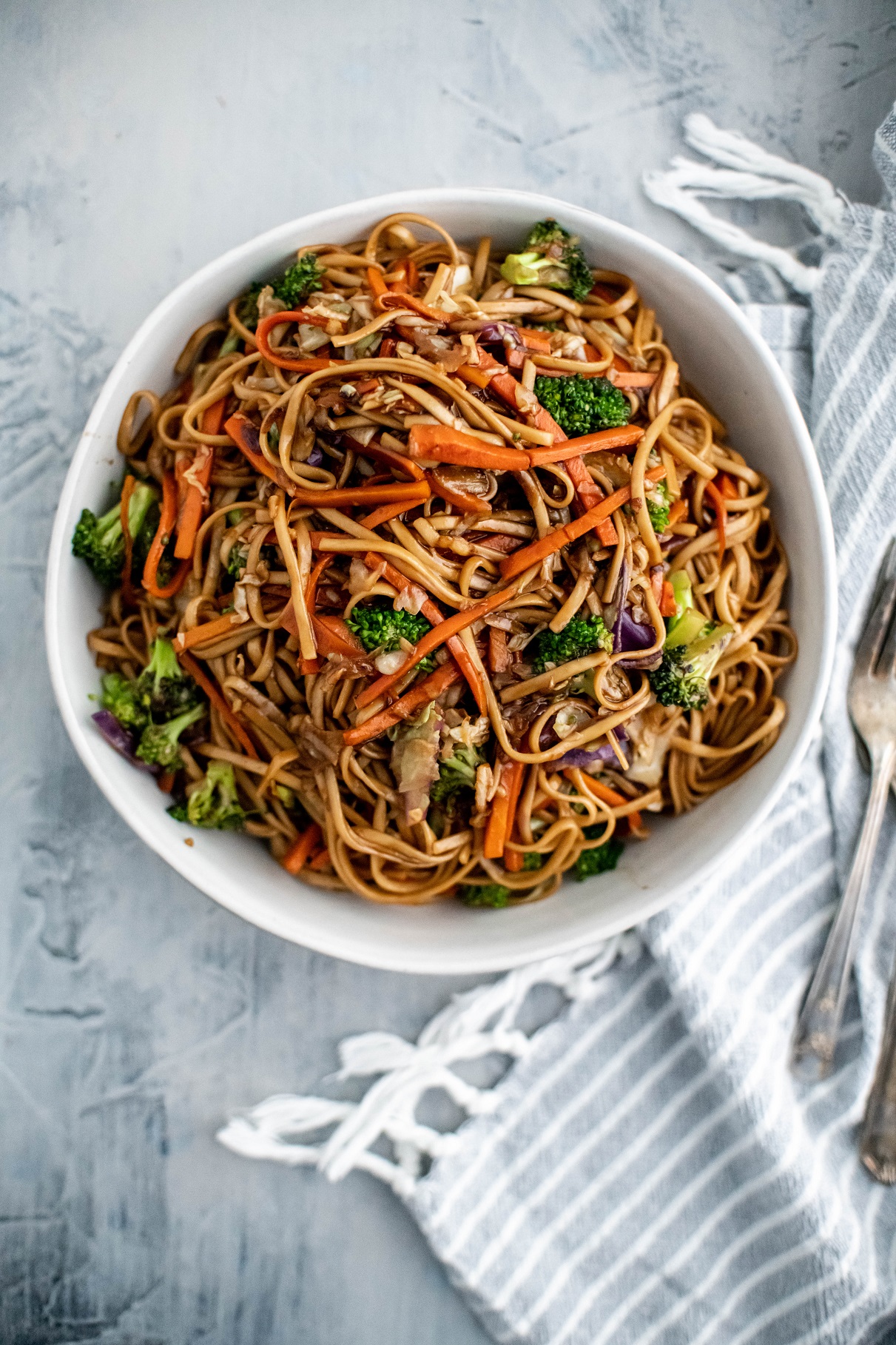 White bowl filled with veggie lo mein. Blue and white striped napkin to the right with a pile of antique forks.