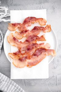 Parcooked bacon on a white round plate lined with paper towels.