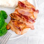 Bacon wrapped pork chop on a white round plate with broccoli and mashed potatoes on the side.