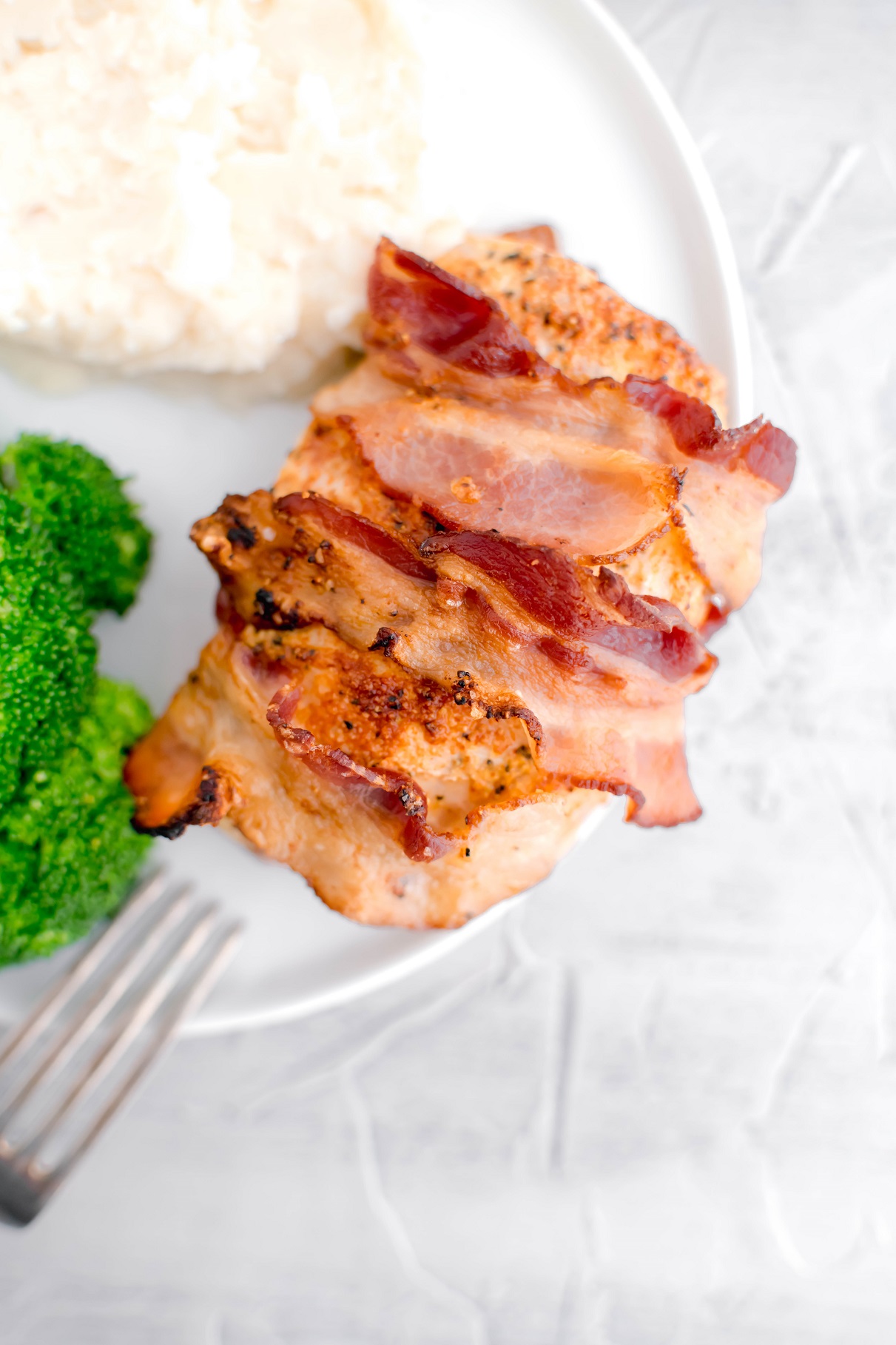 Bacon wrapped pork chop on a white round plate with broccoli and mashed potatoes on the side.