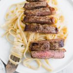 Fettucine alfredo on a plate topped with sliced steak.