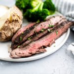 Thin slices of flank steak on a white round plate with a baked potato and steamed broccoli.