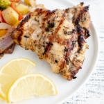 Two grilled lemon herb chicken thighs on a plate with grilled vegetables.