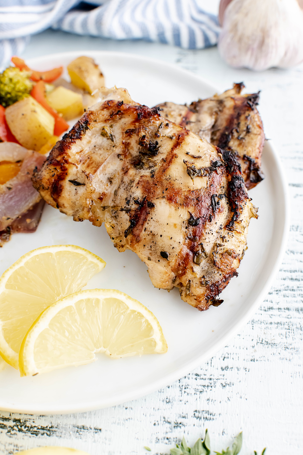 Two grilled lemon herb chicken thighs on a plate with grilled vegetables.