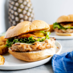 Teriyaki chicken burger on a round white plate with tater tots on the side. Fresh pineapple in the background.