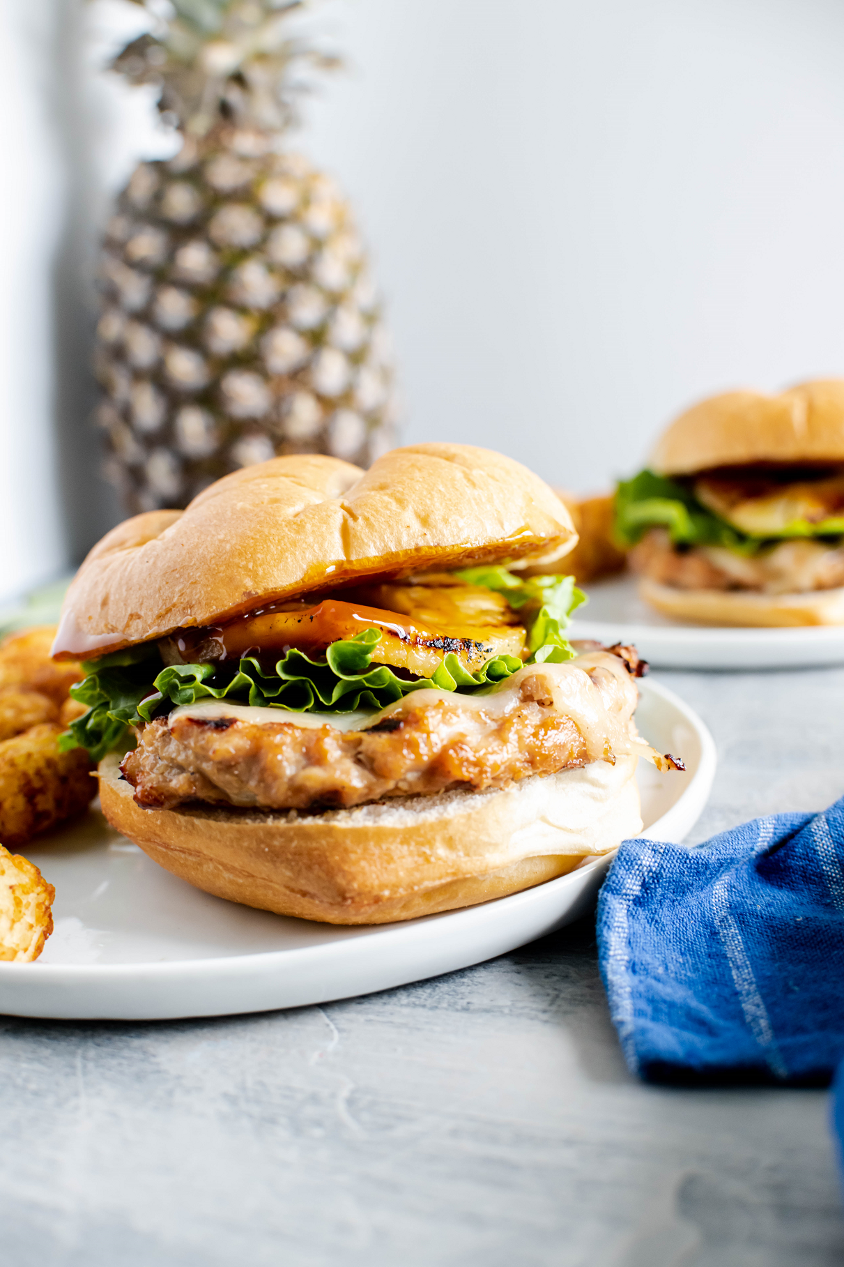 Teriyaki chicken burger on a round white plate with tater tots on the side. Fresh pineapple in the background.