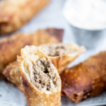 Tray covered in newspaper printed parchment. Several cheeseburger egg rolls on tray along with one cut on a diagonal to show interior.