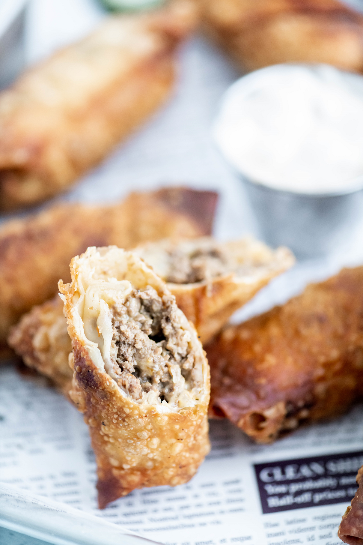 Tray covered in newspaper printed parchment. Several cheeseburger egg rolls on tray along with one cut on a diagonal to show interior.