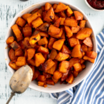 Cream colored bowl filled with diced roasted sweet potatoes. Bottle of pure maple syrup in the background.
