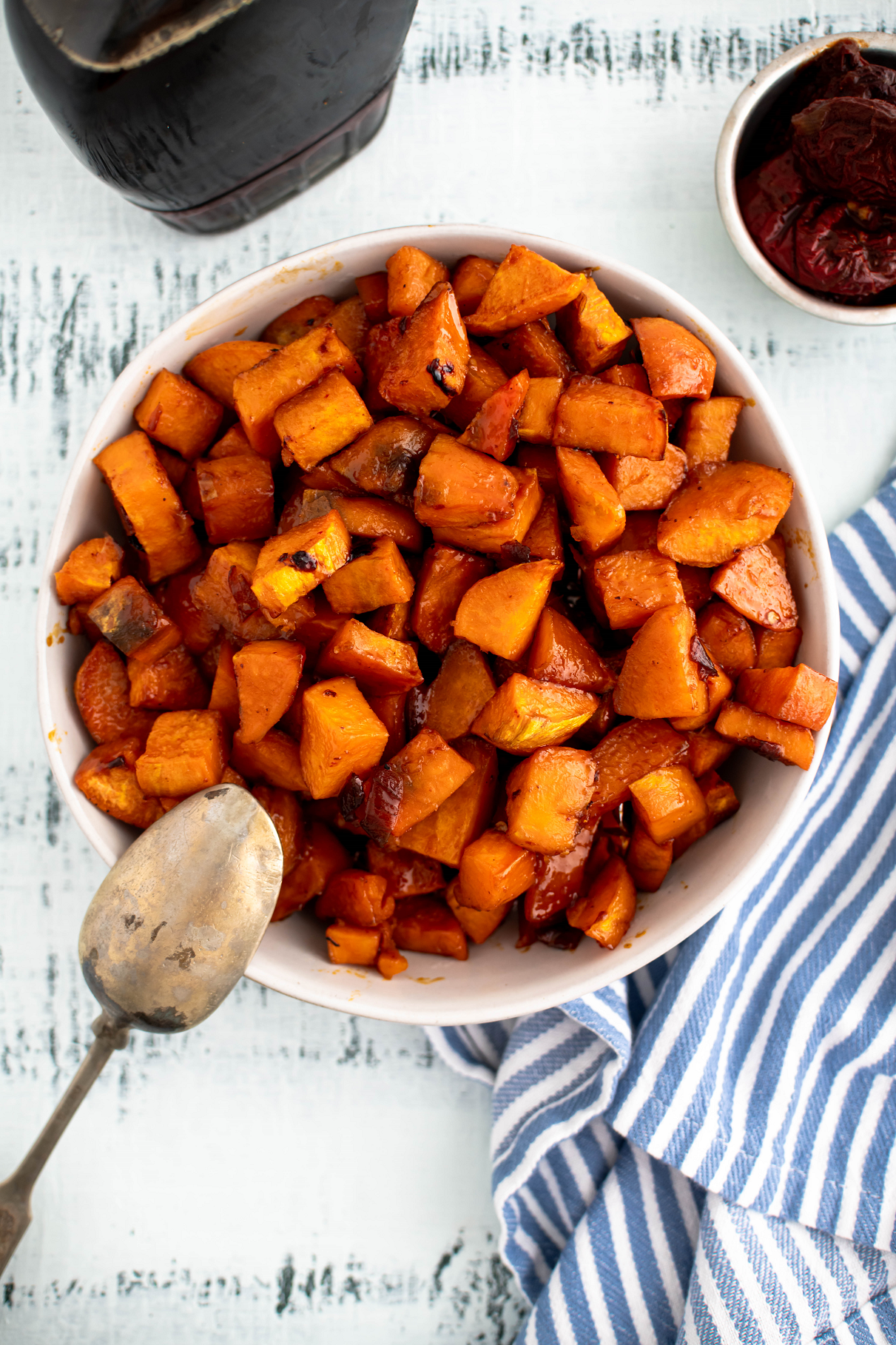 Cream colored bowl filled with diced roasted sweet potatoes. Bottle of pure maple syrup in the background.