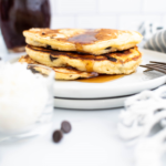 Stack of three pancakes on a round white plate with a glass bottle of maple syrup in the background.