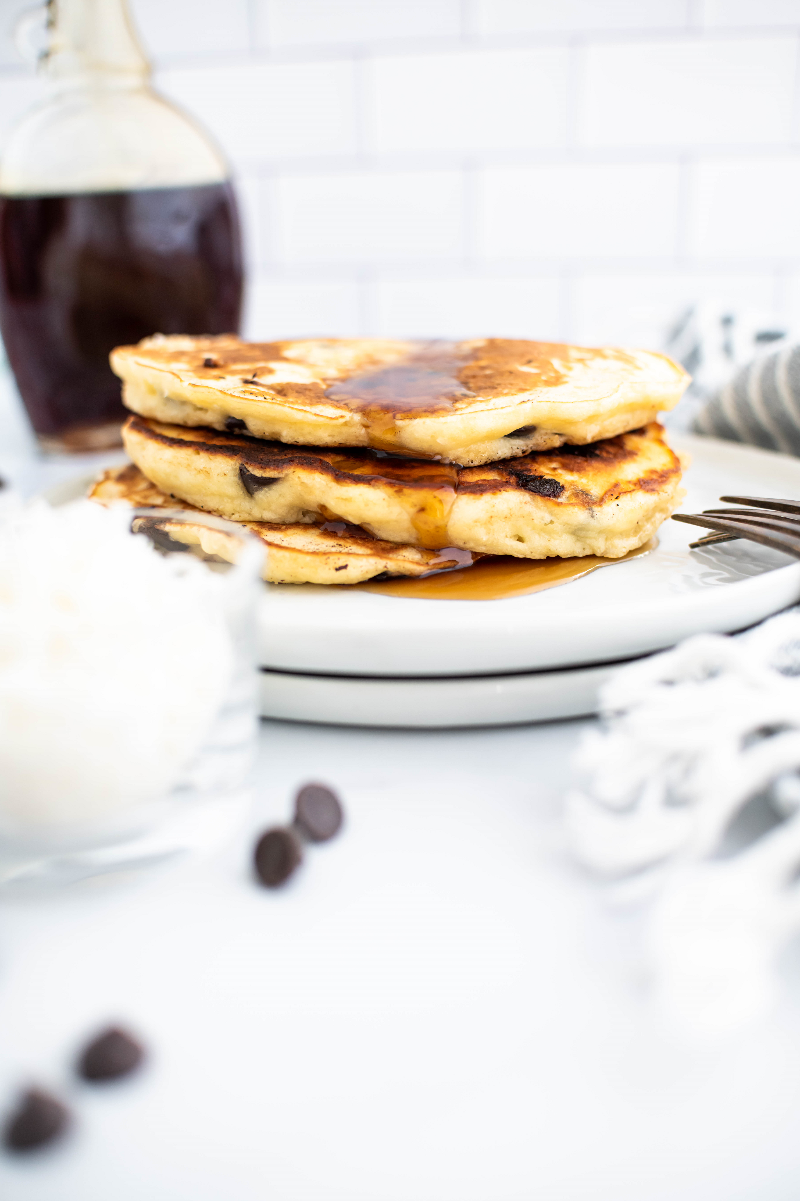 Stack of three pancakes on a round white plate with a glass bottle of maple syrup in the background.