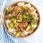 Brown bowl filled with white rice and topped with ground pork and broccoli stir fry. Blue and white striped towel on the left side along with a set of chopsticks.