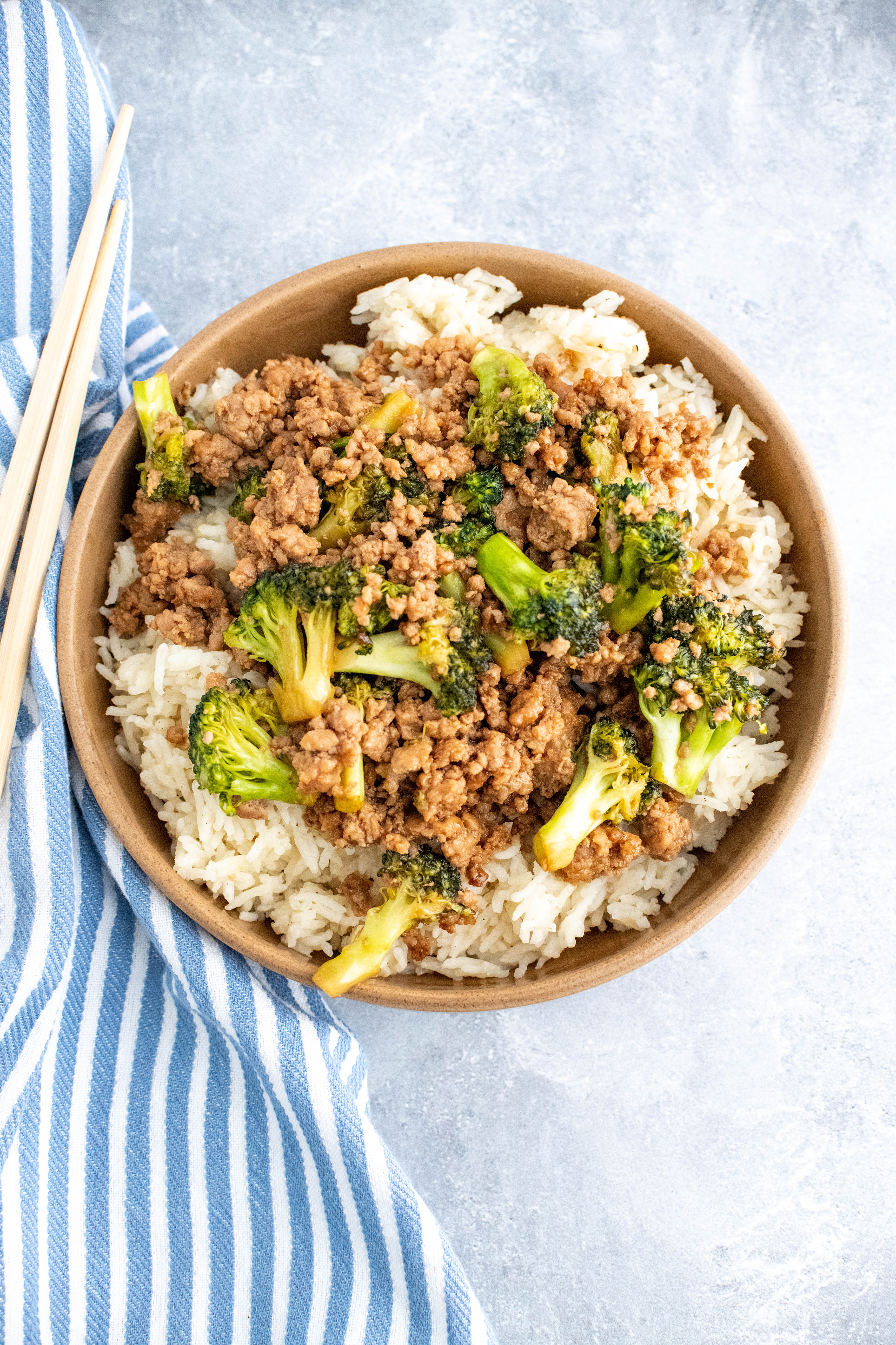 Brown bowl filled with white rice and topped with ground pork and broccoli stir fry. Blue and white striped towel on the left side along with a set of chopsticks.