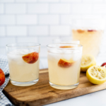 Two glasses of peach lemonade on a wood cutting board with a pitcher of lemonade in the background.