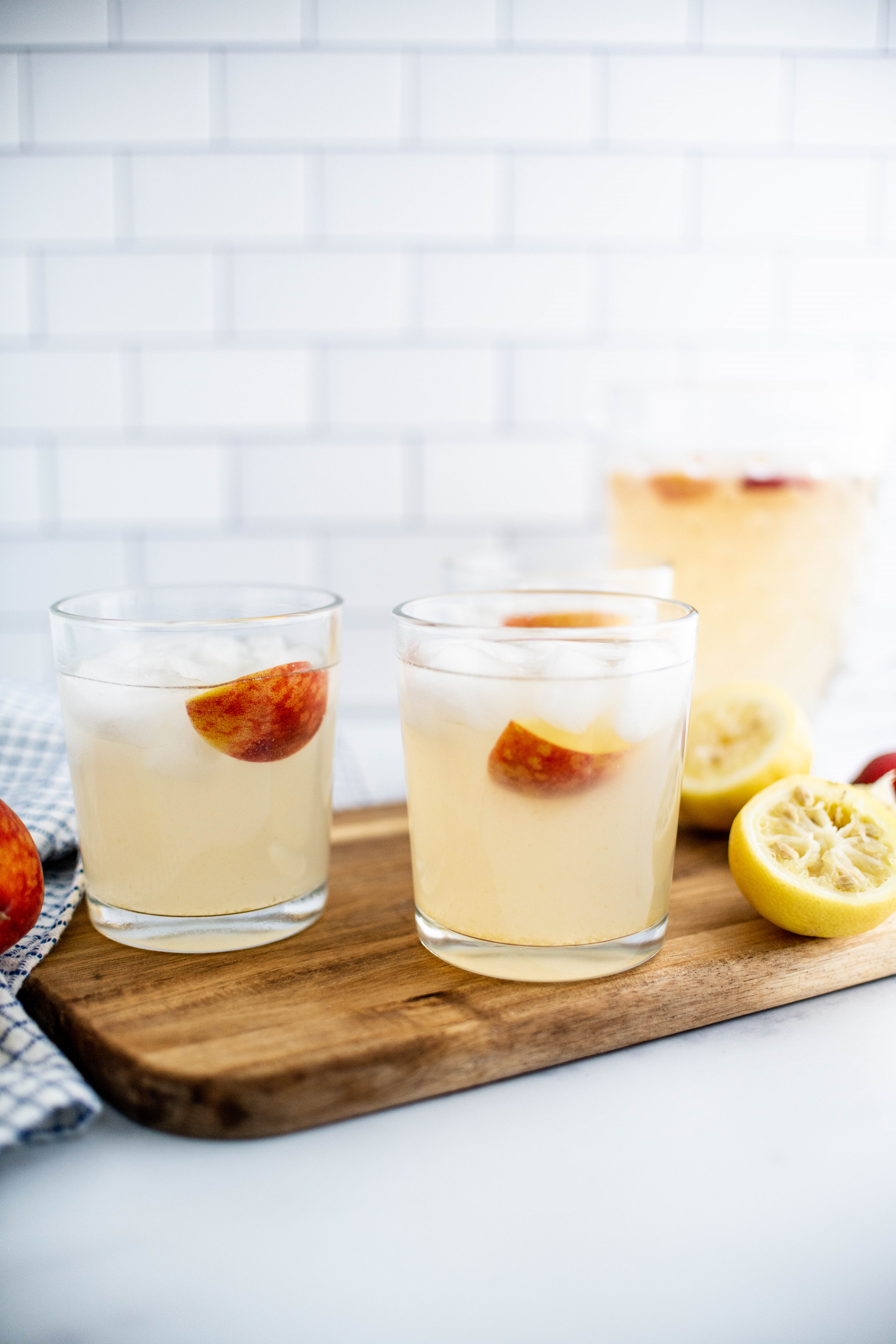 Two glasses of peach lemonade on a wood cutting board with a pitcher of lemonade in the background.