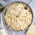 Gray bowl filled with pesto alfredo. Fork twirled with pasta in the bowl. Microplane and chunk of parmesan in the bottom corner and bottle of heavy cream in the upper corner.