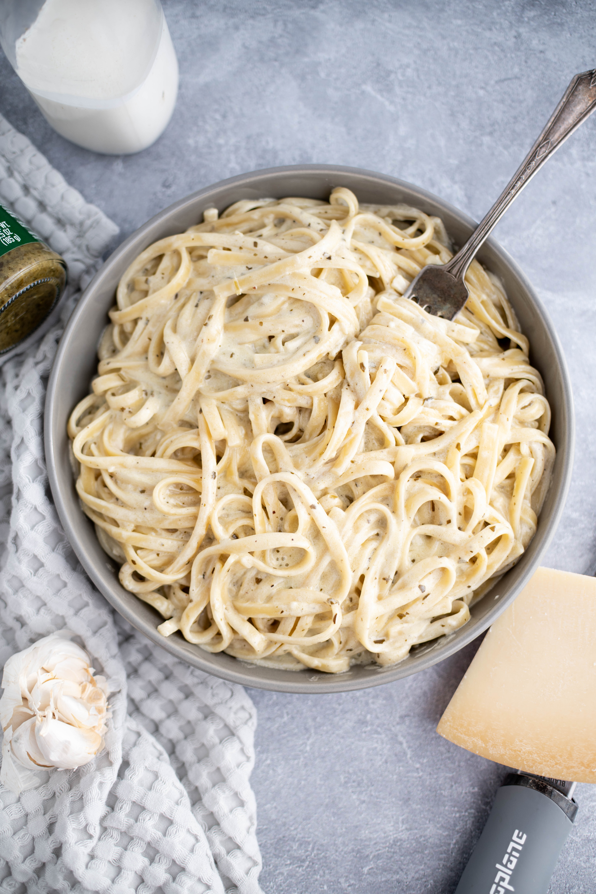 Gray bowl filled with pesto alfredo. Fork twirled with pasta in the bowl. Microplane and chunk of parmesan in the bottom corner and bottle of heavy cream in the upper corner.