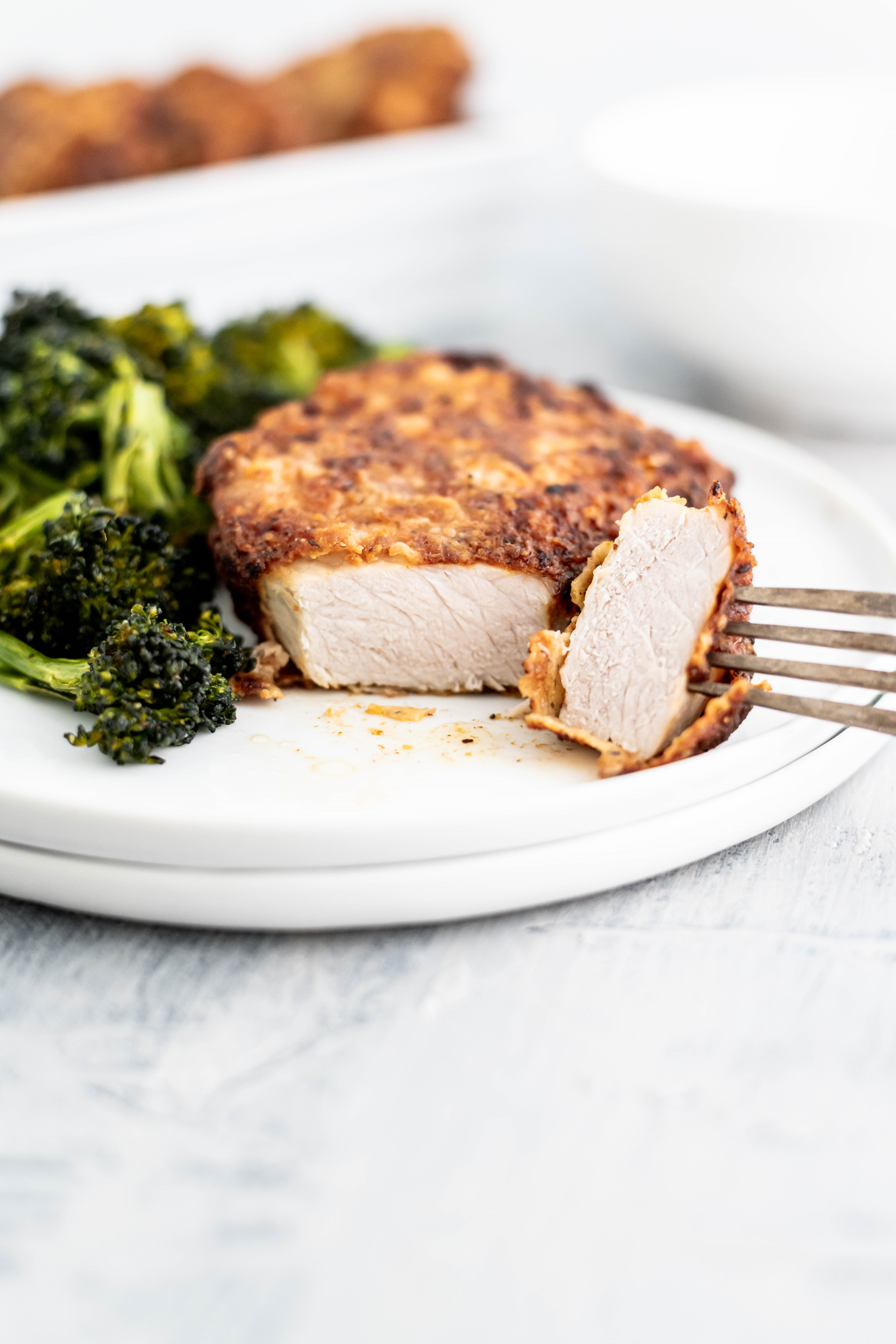 Southern Fried Pork Chop on a round white plate with a bite cut off and on the fork to show the interior of the pork chop. Roasted broccoli on the side.