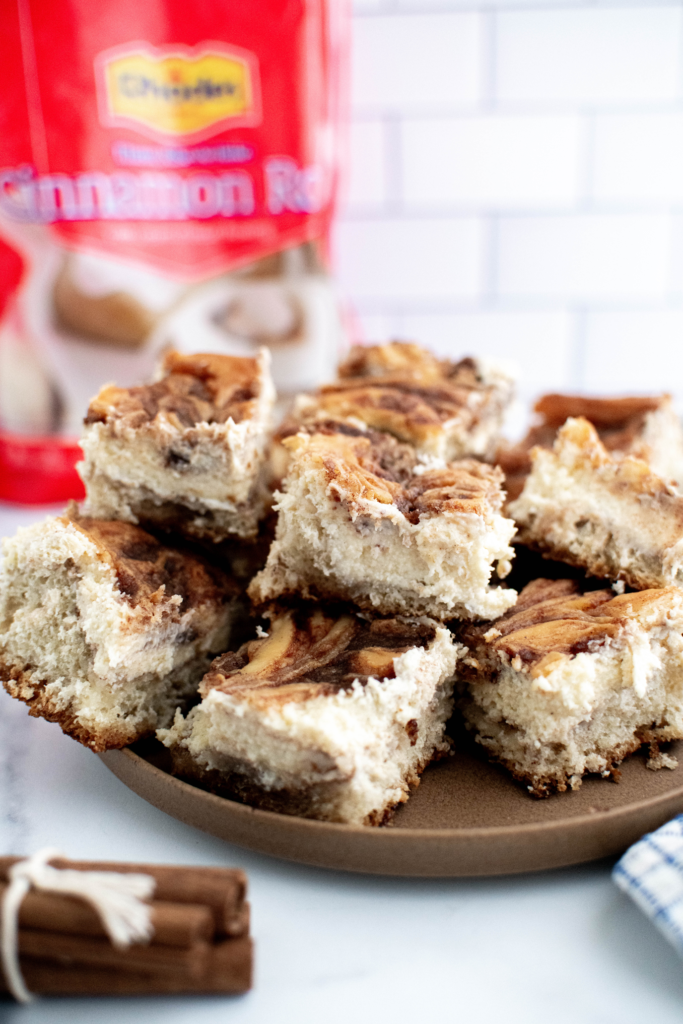 Pile of cinnamon roll cheesecake bars on a round sand colored plate with a bag of Rhodes cinnamon rolls in the background
