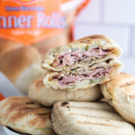 Grilled Cuban Sandwich Pockets in a pile on a roun white plate with one cut in half to show the interior. Bag of Rhodes bread rolls in the background.