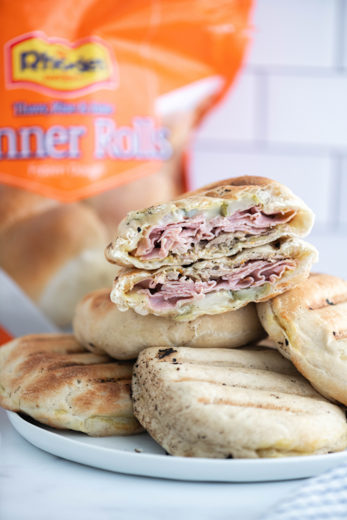 Grilled Cuban Sandwich Pockets in a pile on a roun white plate with one cut in half to show the interior. Bag of Rhodes bread rolls in the background.