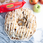 Cinnamon roll apple pie monkey bread on a white hexagon platter with apples and cinnamon roll packaging behind it.