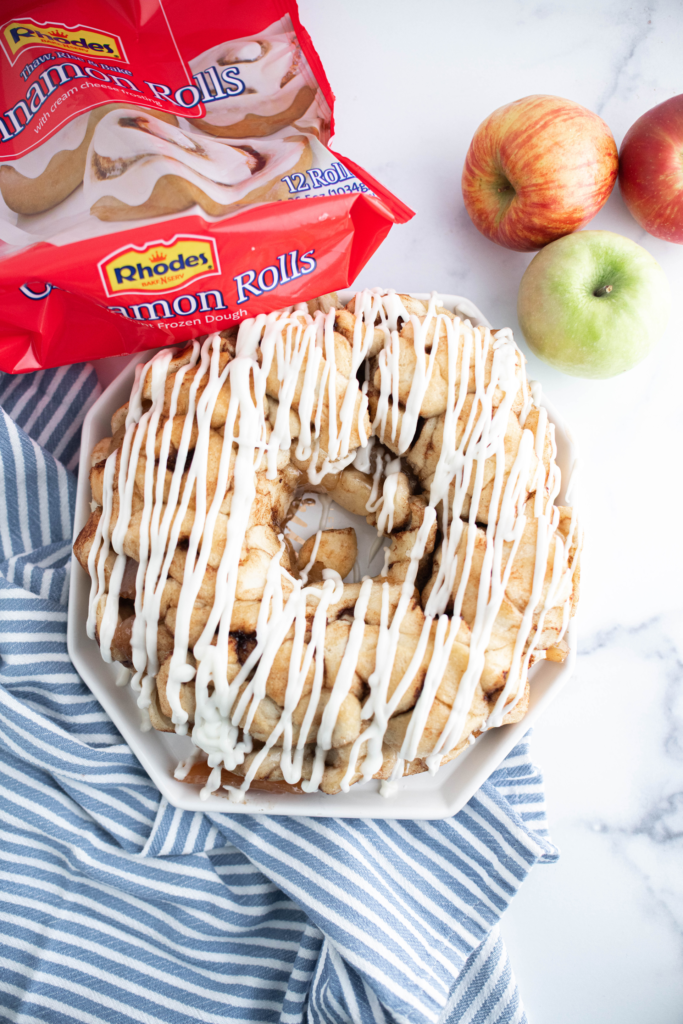 Cinnamon roll apple pie monkey bread on a white hexagon platter with apples and cinnamon roll packaging behind it.
