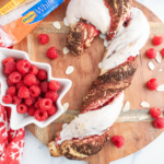 Braided Candy Cane Bread filled with raspberry preserves and sliced almonds, topped with white glaze. Candy Cane bread sitting on a round wooden board.